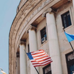 关闭-up view of a neoclassical government building with large columns 和 a facade made of stone. 几个旗帜, 包括美国国旗, are visible in front of the building against a clear blue sky.